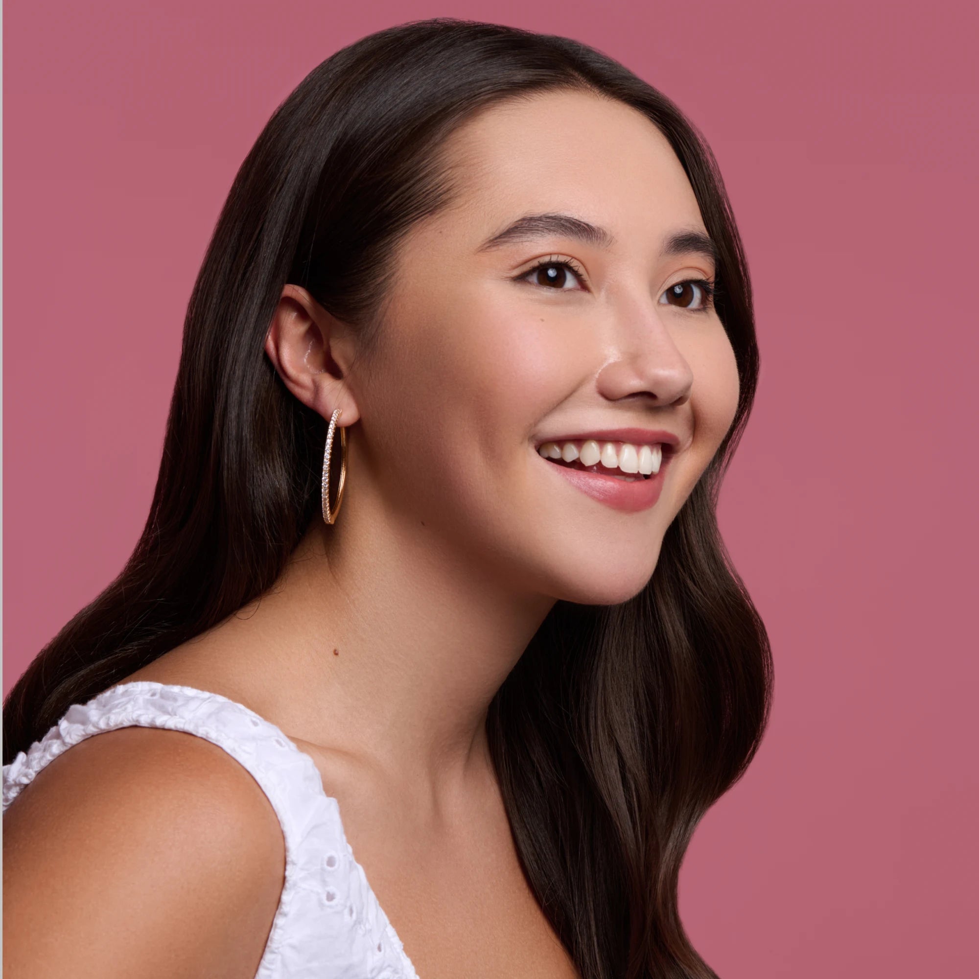 Woman with long dark hair wearing gold hoop earrings against a pink background
