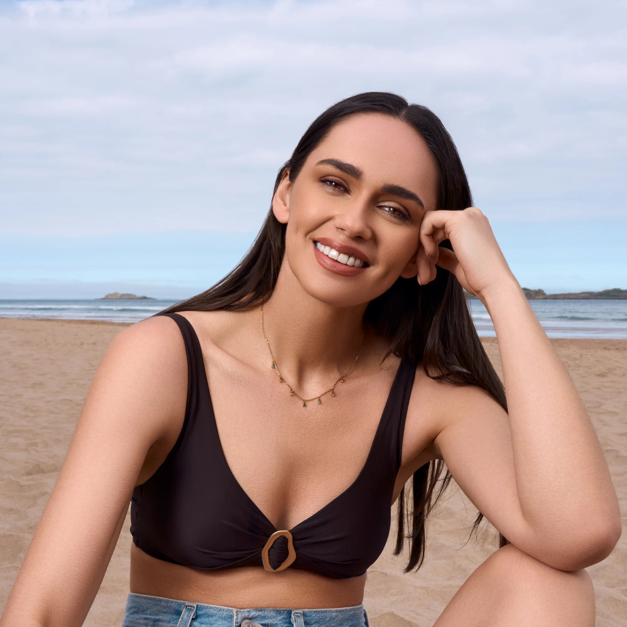 A woman with long dark hair sits on a sandy beach wearing a black bikini top with a front knot detail and a gold-tone ring accent, paired with light blue denim bottoms. The background shows the ocean and a cloudy sky.