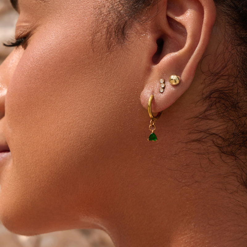 Close-up of an ear with gold hoop earrings featuring green gemstones.